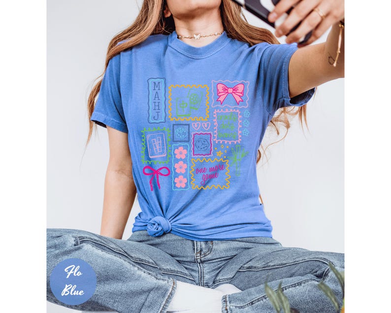 May include: A blue t-shirt with a graphic design featuring various postage stamp-like images. The stamps include a pink bow, flowers, and the text "one more game". The shirt is tied at the waist.