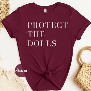 May include: A maroon t-shirt with the text "PROTECT THE DOLLS" printed in white.