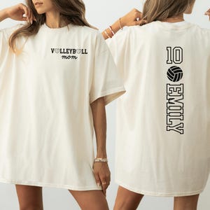 May include: Cream-colored t-shirt with black text. The front reads "VOLLEYBALL mom" above the chest. The back features the number "10", a volleyball graphic, and the name "EMILY" in a vertical layout.