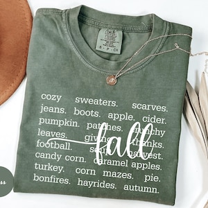 May include: A moss green Comfort Colors t-shirt with the word "fall" in a cursive font. The shirt lists fall-themed words like "cozy," "sweaters," "pumpkin," and "bonfires." A gold necklace with a pendant is on the shirt.