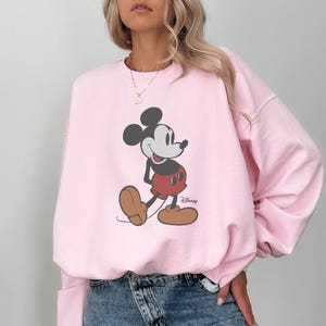 May include: A pale pink sweatshirt with a Mickey Mouse graphic. The cartoon character is in black, red, and brown, with the Disney logo. The sweatshirt is a casual style, suitable for everyday wear.