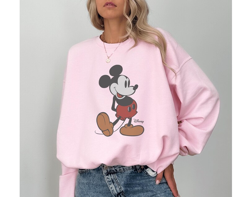 May include: A pale pink sweatshirt with a Mickey Mouse graphic. The cartoon character is in black, red, and brown, with the Disney logo. The sweatshirt is a casual style, suitable for everyday wear.
