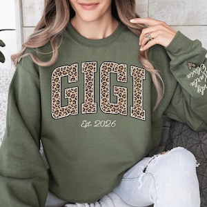 May include: An olive green sweatshirt with the word "GIGI" in large, leopard print letters. Below the word "GIGI" is the text "Est. 2026". The sleeve has the names "Cindy, Karla, Olivia" embroidered on it.