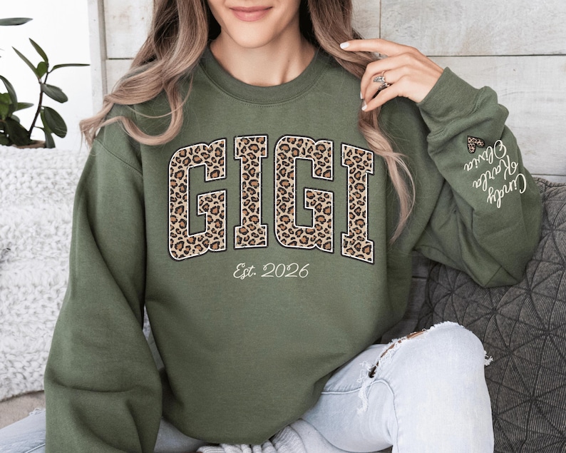 May include: An olive green sweatshirt with the word "GIGI" in large, leopard print letters. Below the word "GIGI" is the text "Est. 2026". The sleeve has the names "Cindy, Karla, Olivia" embroidered on it.