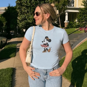 Pu&ograve; includere: T-shirt azzurra con un disegno di Minnie Mouse. Il personaggio dei cartoni animati &egrave; raffigurato in una posa classica, con una gonna a pois e un cappello. La maglietta &egrave; abbinata a jeans blu.