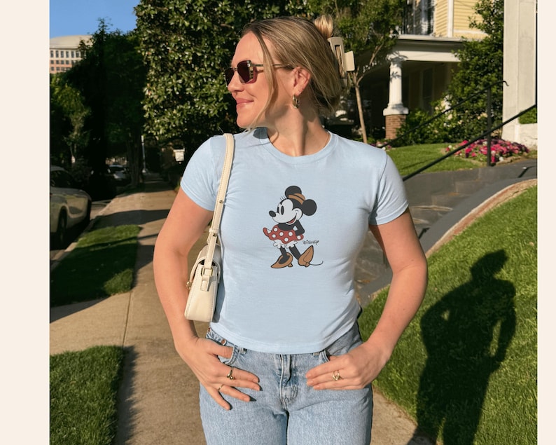 Pu&ograve; includere: T-shirt azzurra con un disegno di Minnie Mouse. Il personaggio dei cartoni animati &egrave; raffigurato in una posa classica, con una gonna a pois e un cappello. La maglietta &egrave; abbinata a jeans blu.