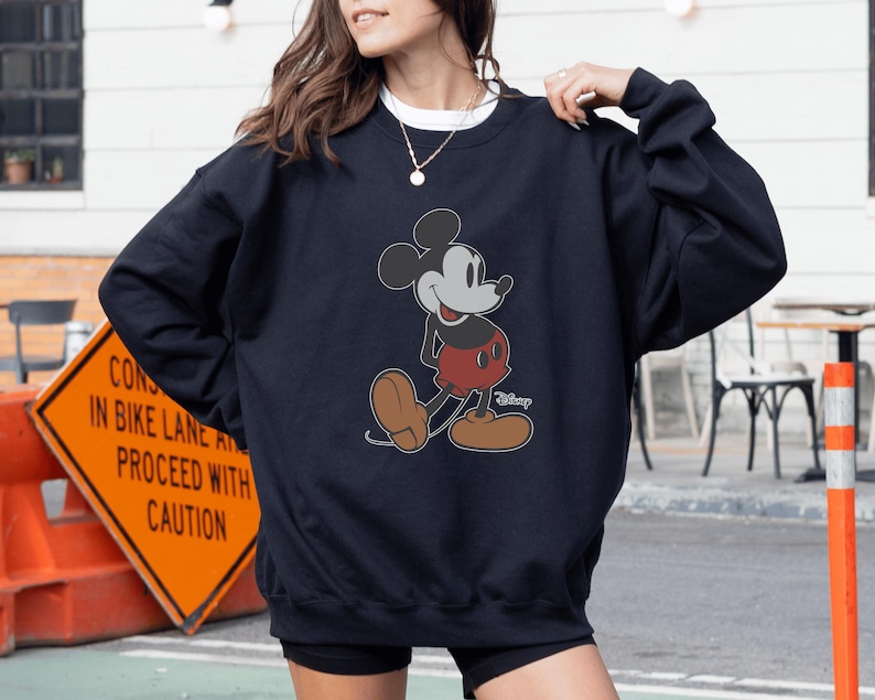 May include: Navy blue sweatshirt featuring a classic Mickey Mouse graphic. The cartoon character is depicted in his iconic pose, wearing red shorts and brown shoes. The sweatshirt is a comfortable, casual style.