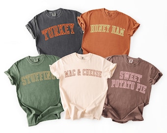 May include: Five short-sleeved t-shirts in various colors, each with a different food-related word in a collegiate font. The shirts are dark gray, orange, green, cream, and brown. The words are "TURKEY", "HONEY HAM", "STUFFING", "MAC & CHEESE", and "SWEET POTATO PIE".