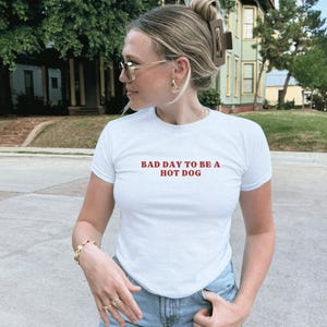 Puede incluir: Camiseta blanca con el texto rojo "BAD DAY TO BE A HOT DOG". La camiseta es de manga corta y tiene cuello redondo. La persona lleva joyas y gafas de sol doradas. El fondo muestra una calle residencial con casas y &aacute;rboles.