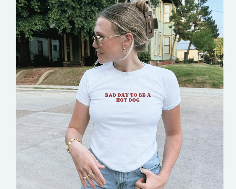 Puede incluir: Camiseta blanca con el texto rojo "BAD DAY TO BE A HOT DOG". La camiseta es de manga corta y tiene cuello redondo. La persona lleva joyas y gafas de sol doradas. El fondo muestra una calle residencial con casas y &aacute;rboles.
