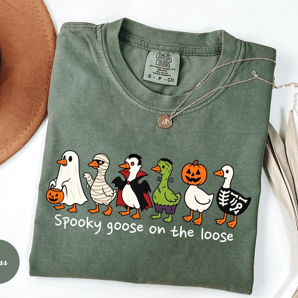 May include: A moss green crew neck t-shirt featuring a graphic of six geese dressed in Halloween costumes, including a ghost, mummy, vampire, Frankenstein monster, pumpkin, and skeleton. The text below reads, "Spooky goose on the loose."