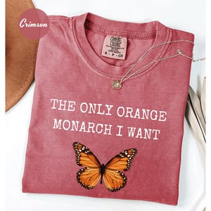 Pode incluir: Camiseta cor carmesim com o texto "THE ONLY ORANGE MONARCH I WANT" e um gr&aacute;fico de borboleta. Um colar de ouro com um pingente repousa sobre a camiseta. A camiseta est&aacute; dobrada sobre uma superf&iacute;cie branca.
