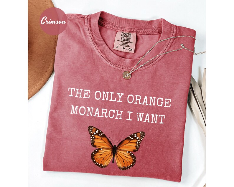 Pode incluir: Camiseta cor carmesim com o texto "THE ONLY ORANGE MONARCH I WANT" e um gr&aacute;fico de borboleta. Um colar de ouro com um pingente repousa sobre a camiseta. A camiseta est&aacute; dobrada sobre uma superf&iacute;cie branca.