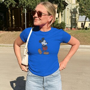 Może przedstawiać: Kr&oacute;lewsko niebieska koszulka z klasyczną grafiką Myszki Miki. Postać z kresk&oacute;wki jest przedstawiona w czerwonych spodenkach, ż&oacute;łtych butach i czarnych rękawicach. Koszulka jest połączona z jasnoniebieskimi szortami dżinsowymi i białą torbą na ramię.