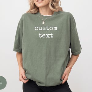 K&ouml;nnte beinhalten: Ein &uuml;bergro&szlig;es, moosgr&uuml;nes T-Shirt mit dem wei&szlig;en Aufdruck "custom text". Das Shirt hat kurze &Auml;rmel und eine lockere Passform. Das Model tr&auml;gt eine schwarze Hose und eine Halskette.