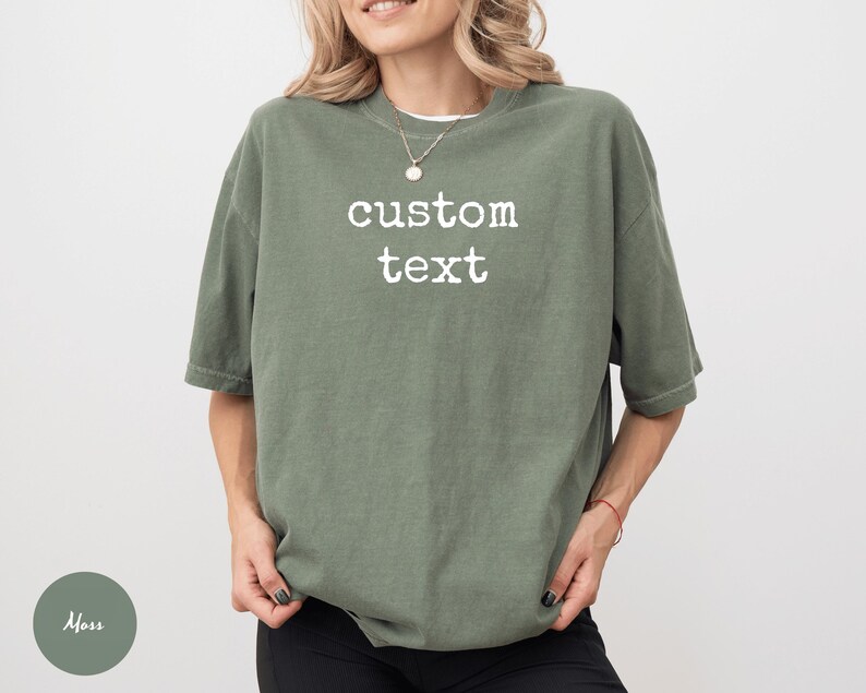 K&ouml;nnte beinhalten: Ein &uuml;bergro&szlig;es, moosgr&uuml;nes T-Shirt mit dem wei&szlig;en Aufdruck "custom text". Das Shirt hat kurze &Auml;rmel und eine lockere Passform. Das Model tr&auml;gt eine schwarze Hose und eine Halskette.