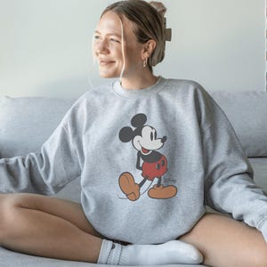 May include: A light gray sweatshirt featuring a classic Mickey Mouse graphic. The cartoon character is depicted in his iconic red shorts, brown shoes, and black gloves. The sweatshirt has a crew neck and long sleeves.