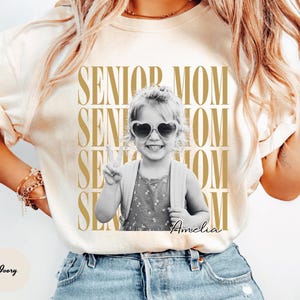 May include: An ivory t-shirt featuring a repeating gold text graphic that reads SENIOR MOM. Centered is a black and white photograph of a child wearing heart-shaped sunglasses and a backpack, making a peace sign. The name Amelia is written below.