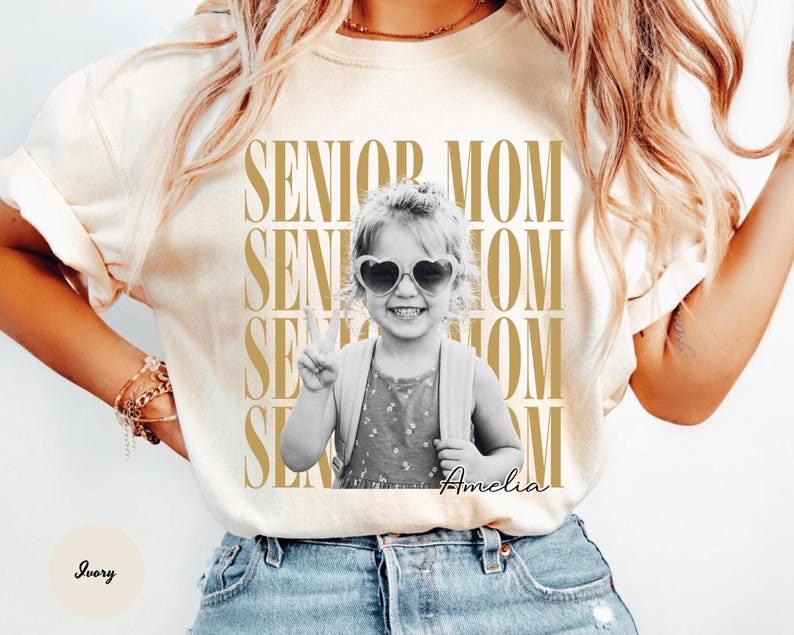 May include: An ivory t-shirt featuring a repeating gold text graphic that reads SENIOR MOM. Centered is a black and white photograph of a child wearing heart-shaped sunglasses and a backpack, making a peace sign. The name Amelia is written below.