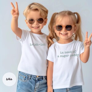 K&ouml;nnte beinhalten: Zwei Kinder mit wei&szlig;en T-Shirts und Sonnenbrillen. Auf einem Shirt steht "irish-ish", auf dem anderen "in need of a sugar paddy". Beide Kinder l&auml;cheln und zeigen das Peace-Zeichen.