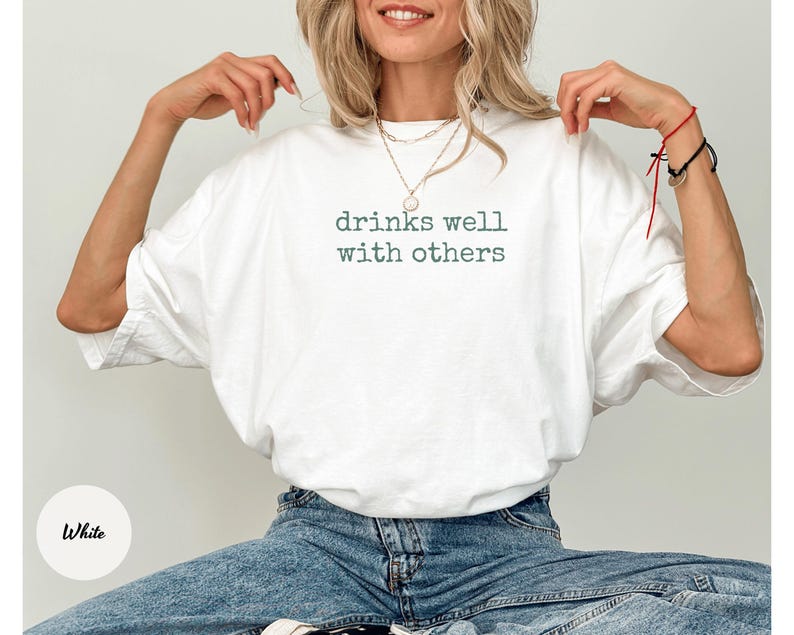 K&ouml;nnte beinhalten: Wei&szlig;es T-Shirt mit dem Text "drinks well with others" in gr&uuml;ner Schrift. Das Shirt hat einen l&auml;ssigen Stil mit lockerer Passform. Das Model tr&auml;gt eine blaue Jeans.