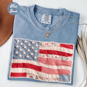 May include: A light blue t-shirt with a patchwork American flag design. The flag features white stars on a blue background and red and floral-patterned stripes. A gold necklace with a pendant rests on the shirt. The text "Blue Jean" is in a circle in the upper left corner.