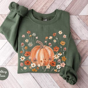 May include: A military green crewneck sweatshirt featuring a central graphic of a large orange pumpkin surrounded by a variety of small, colorful wildflowers in shades of red, orange, yellow, and white, with a small circular label reading Military Green.