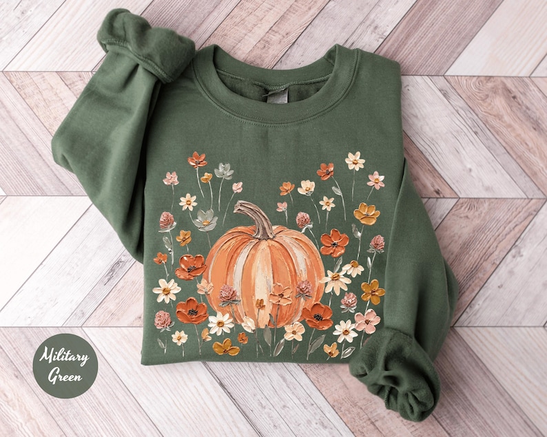 May include: A military green crewneck sweatshirt featuring a central graphic of a large orange pumpkin surrounded by a variety of small, colorful wildflowers in shades of red, orange, yellow, and white, with a small circular label reading Military Green.