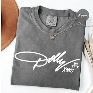 May include: A pepper-colored t-shirt featuring the white script text Dolly XOXO with three small heart outlines. The shirt has a crew neckline and a Comfort Colors brand label on the inside collar showing size S-P-CH and 100% cotton material.