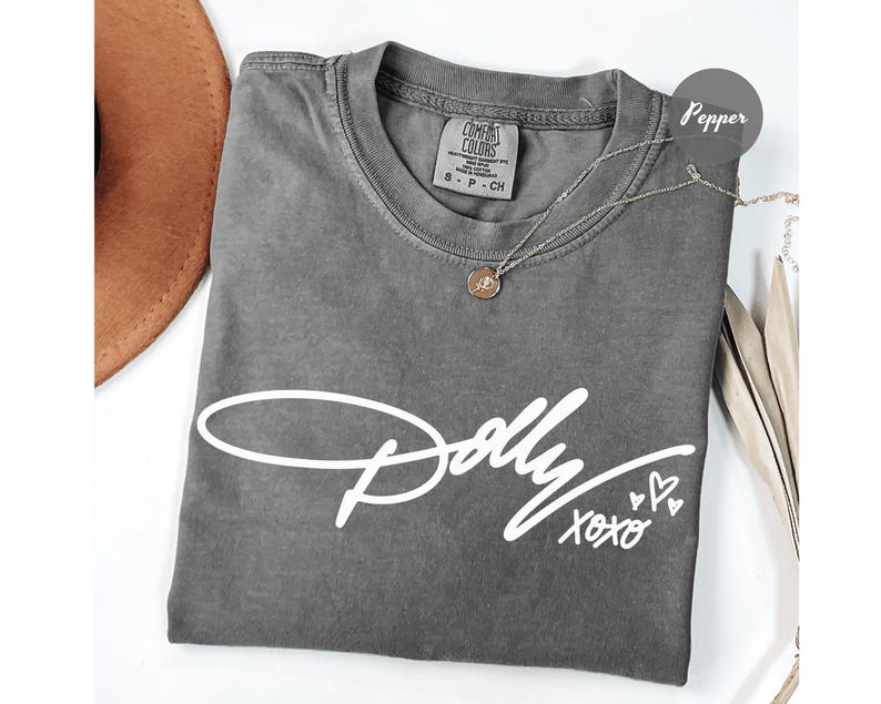 May include: A pepper-colored t-shirt featuring the white script text Dolly XOXO with three small heart outlines. The shirt has a crew neckline and a Comfort Colors brand label on the inside collar showing size S-P-CH and 100% cotton material.