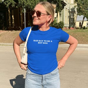 Puede incluir: Una camiseta azul real con el texto blanco "BAD DAY TO BE A HOT DOG". La camiseta se combina con vaqueros de mezclilla de lavado claro. Se lleva un bolso blanco al hombro. La persona lleva gafas de sol.