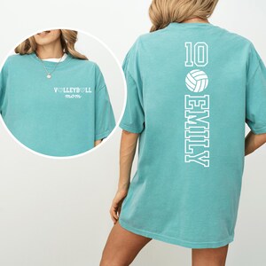 May include: A teal-colored t-shirt with the text "VOLLEYBALL mom" on the front. The back of the shirt features the number 10, a volleyball graphic, and the name "EMILY" in white lettering. The shirt has short sleeves.