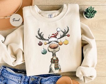 Op de afbeelding: Een zandkleurige sweatshirt met een grillig rendierontwerp. Het rendier draagt een kerstmuts, een groene en rode sjaal en heeft ballen aan zijn gewei hangen. De sweatshirt is een gezellig, feestelijk kledingstuk.