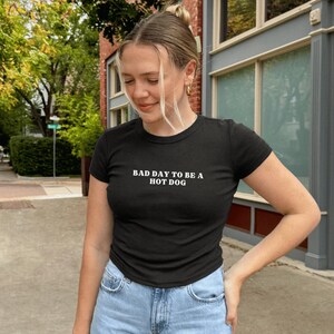 Puede incluir: Camiseta negra de cuello redondo con el texto blanco "BAD DAY TO BE A HOT DOG". La camiseta se combina con vaqueros de mezclilla azul claro. La imagen est&aacute; tomada al aire libre con un fondo borroso de una calle y edificios.