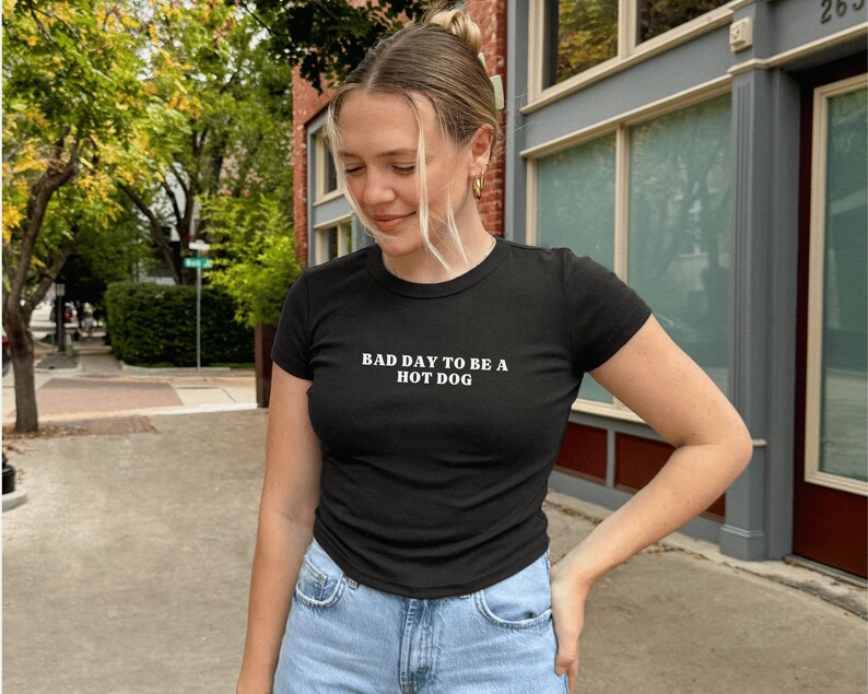 Puede incluir: Camiseta negra de cuello redondo con el texto blanco "BAD DAY TO BE A HOT DOG". La camiseta se combina con vaqueros de mezclilla azul claro. La imagen est&aacute; tomada al aire libre con un fondo borroso de una calle y edificios.