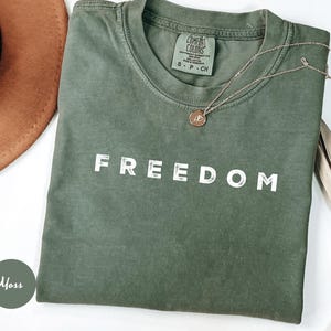 May include: A moss green t-shirt with the word "FREEDOM" printed in white. A gold necklace with a pendant rests on the shirt. A brown hat is partially visible in the upper left corner. The shirt is labeled "Comfort Colors".