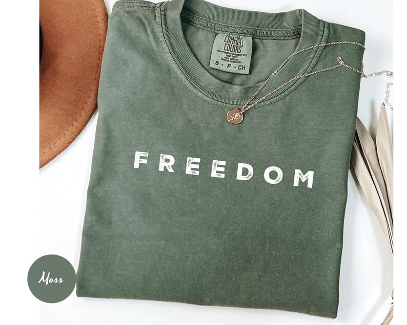 May include: A moss green t-shirt with the word "FREEDOM" printed in white. A gold necklace with a pendant rests on the shirt. A brown hat is partially visible in the upper left corner. The shirt is labeled "Comfort Colors".