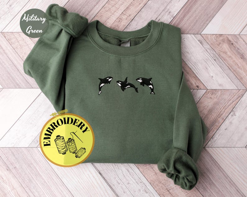 May include: A military green sweatshirt with embroidered orca whales. The sweatshirt features three black and white orcas in a row. The words "Military Green" are in a circle on the top left. The word "EMBROIDERY" is in a circle on the bottom left.