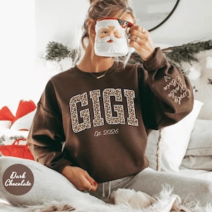 May include: Dark chocolate brown sweatshirt with the word "GIGI" in a leopard print design. The text "Est. 2026" is below the word "GIGI". The sleeve has the names "Cindy, Karla, Olivia" embroidered on it.