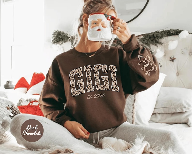 May include: Dark chocolate brown sweatshirt with the word "GIGI" in a leopard print design. The text "Est. 2026" is below the word "GIGI". The sleeve has the names "Cindy, Karla, Olivia" embroidered on it.