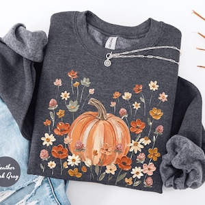 May include: A heather dark gray crewneck sweatshirt featuring a central graphic of a large orange pumpkin surrounded by a variety of small, colorful painted flowers in shades of orange, white, and pink. The Gildan brand tag is visible at the neck.