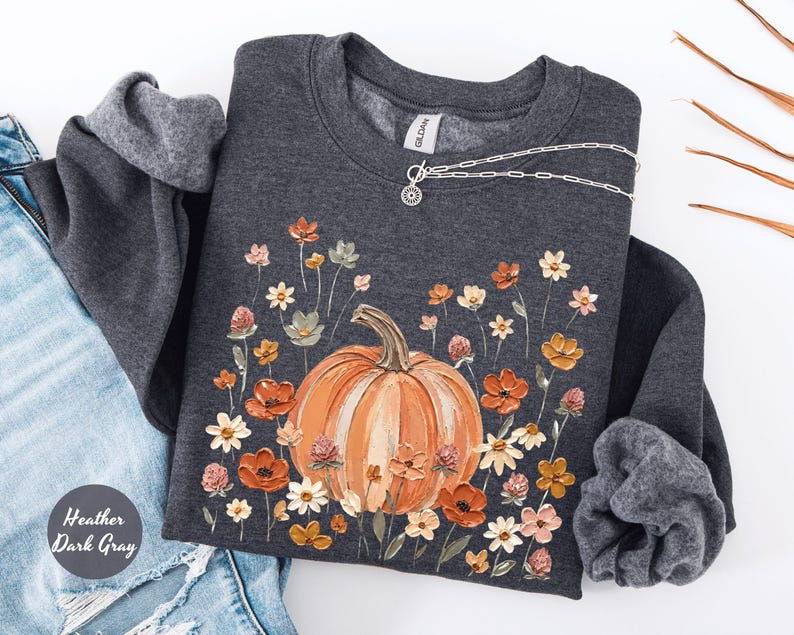 May include: A heather dark gray crewneck sweatshirt featuring a central graphic of a large orange pumpkin surrounded by a variety of small, colorful painted flowers in shades of orange, white, and pink. The Gildan brand tag is visible at the neck.