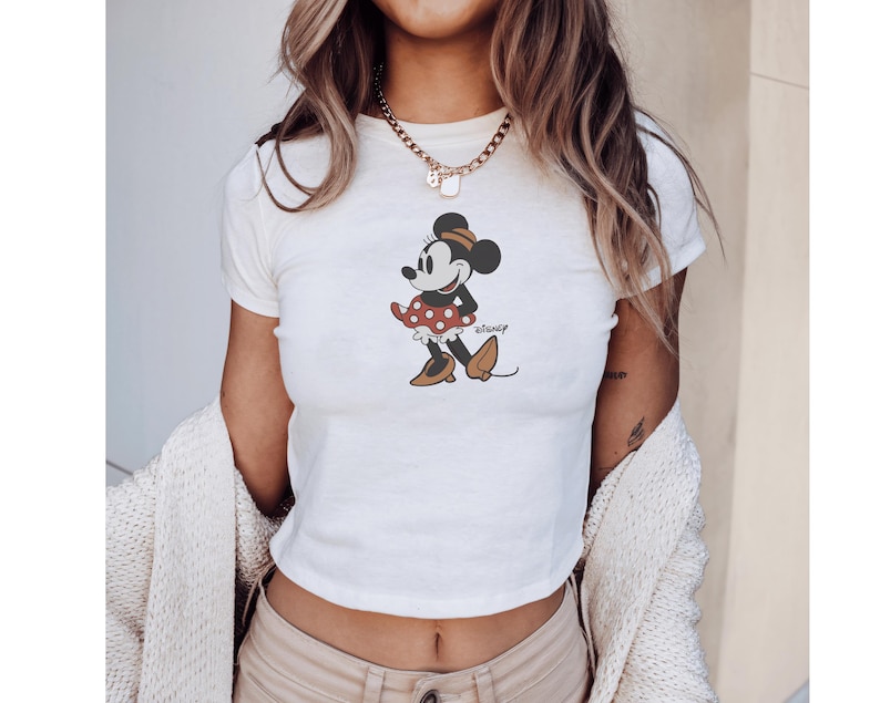 Pu&ograve; includere: Top corto bianco con grafica di Minnie Mouse. Il personaggio dei cartoni animati &egrave; raffigurato in una posa classica, con una gonna a pois rossi e scarpe marroni. Il logo Disney &egrave; stampato sotto.
