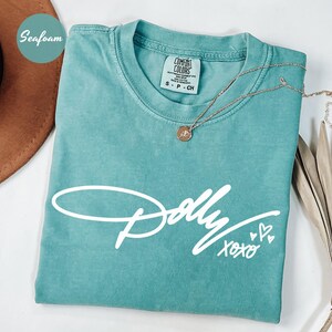 May include: A seafoam green short-sleeve t-shirt featuring the white script text Dolly XOXO with two small heart icons. The shirt has a crew neckline and a Comfort Colors brand label on the inside collar indicating a size small.