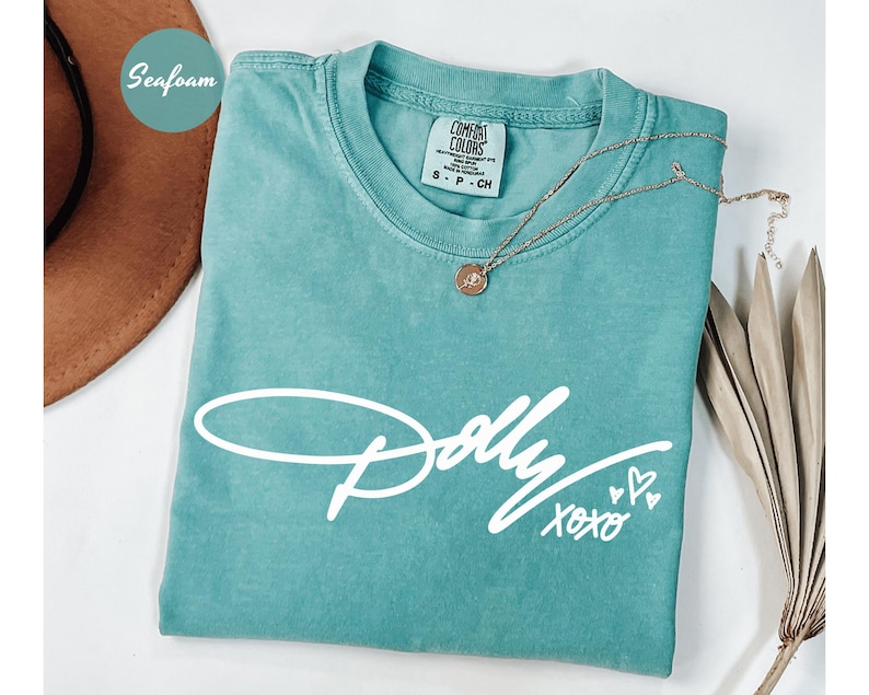 May include: A seafoam green short-sleeve t-shirt featuring the white script text Dolly XOXO with two small heart icons. The shirt has a crew neckline and a Comfort Colors brand label on the inside collar indicating a size small.