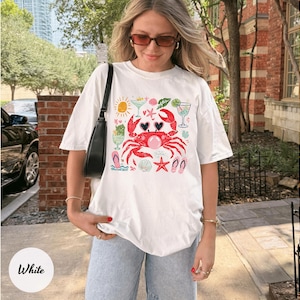 May include: White t-shirt featuring a red crab with heart-shaped sunglasses, surrounded by beach-themed illustrations. The design includes a sun, cocktails, seashells, and flip-flops, perfect for a summer vibe.