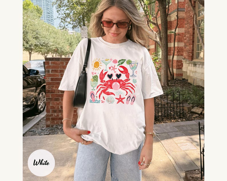May include: White t-shirt featuring a red crab with heart-shaped sunglasses, surrounded by beach-themed illustrations. The design includes a sun, cocktails, seashells, and flip-flops, perfect for a summer vibe.