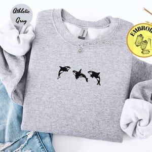May include: Athletic gray sweatshirt with three black and white orca whale embroidery. The sweatshirt has a crew neck and long sleeves. The text "Athletic Gray" is in a circle on the upper left. A silver necklace with a pendant is on the neckline.