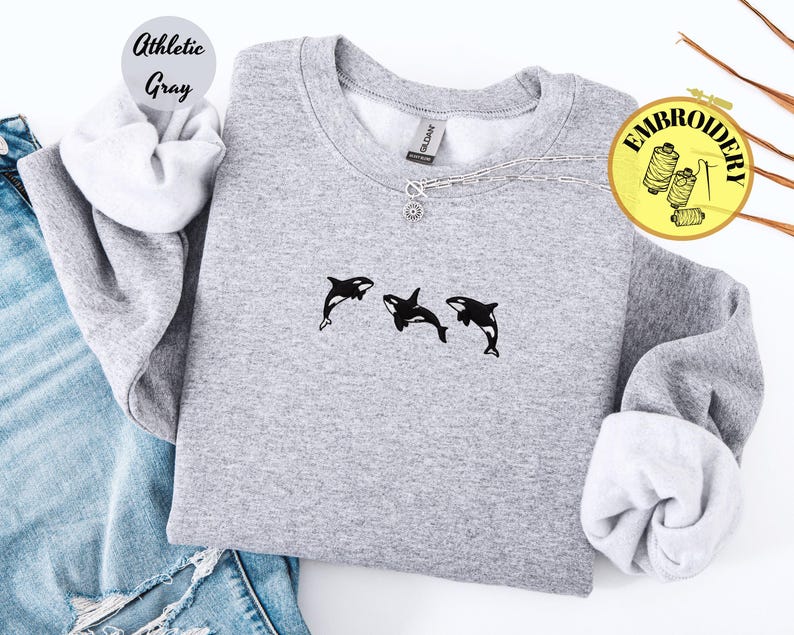May include: Athletic gray sweatshirt with three black and white orca whale embroidery. The sweatshirt has a crew neck and long sleeves. The text "Athletic Gray" is in a circle on the upper left. A silver necklace with a pendant is on the neckline.