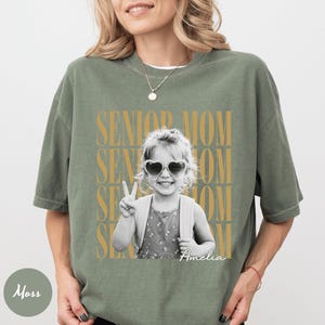 May include: A moss green short-sleeve t-shirt featuring a black and white photograph of a child wearing heart-shaped sunglasses and making a peace sign. The text SENIOR MOM is repeated in gold behind the photo, with the name Amelia in white script.